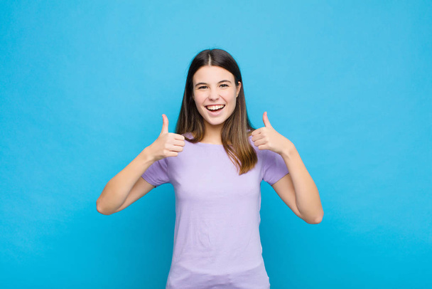 young pretty woman smiling broadly looking happy, positive, confident and successful, with both thumbs up against blue wall - Foto, Bild