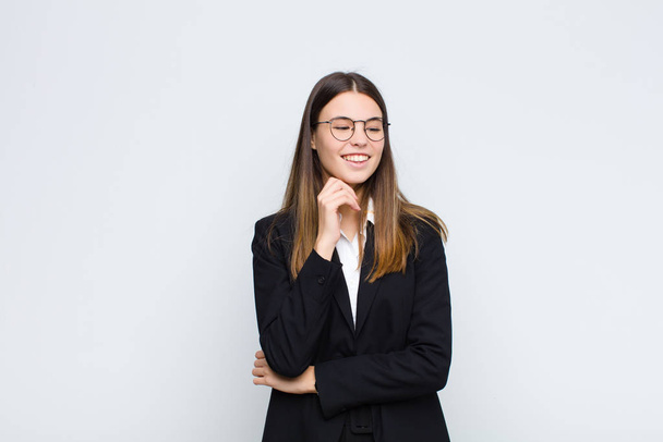 young businesswoman smiling with a happy, confident expression with hand on chin, wondering and looking to the side against white wall - Photo, image