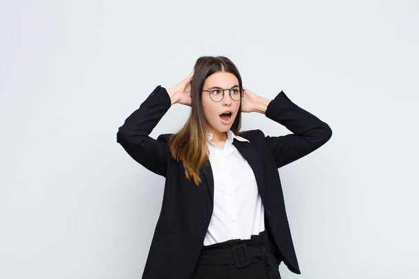young businesswoman with open mouth, looking horrified and shocked because of a terrible mistake, raising hands to head against white wall - Foto, imagen