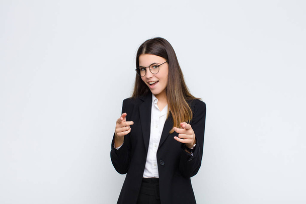 young businesswoman smiling with a positive, successful, happy attitude pointing to the camera, making gun sign with hands against white wall - Foto, Imagen