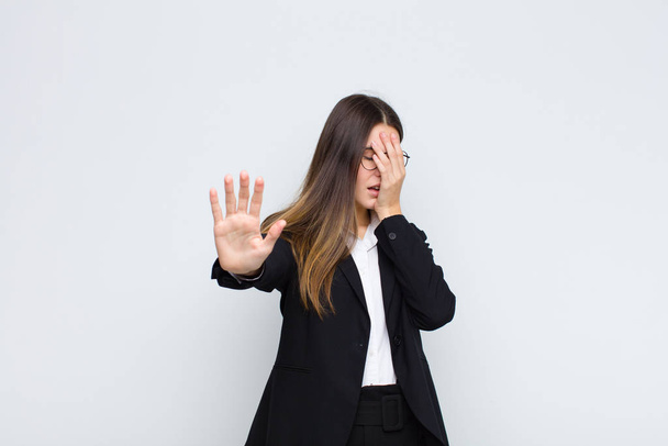young businesswoman covering face with hand and putting other hand up front to stop camera, refusing photos or pictures against white wall - Foto, Bild