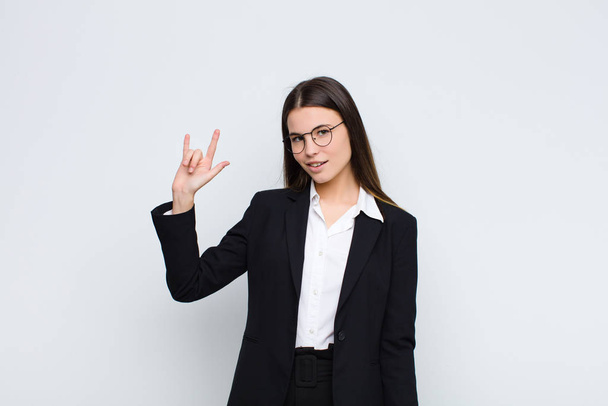 young businesswoman feeling happy, fun, confident, positive and rebellious, making rock or heavy metal sign with hand against white wall - Photo, Image