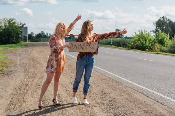 Two beautiful girls hitchhiking and vote with a sign ANYWHERE on road. Copy space. - Fotografie, Obrázek