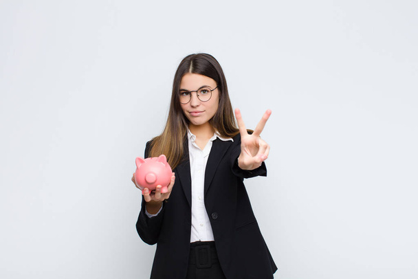 young pretty woman smiling and looking happy, carefree and positive, gesturing victory or peace with one hand with a piggy bank - Photo, Image