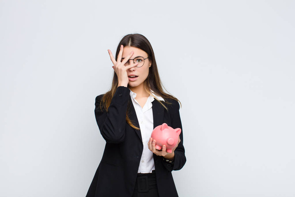 young pretty woman looking shocked, scared or terrified, covering face with hand and peeking between fingers with a piggy bank - Foto, Bild