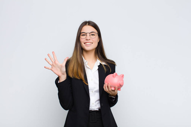 young pretty woman smiling and looking friendly, showing number five or fifth with hand forward, counting down with a piggy bank - Foto, Imagen