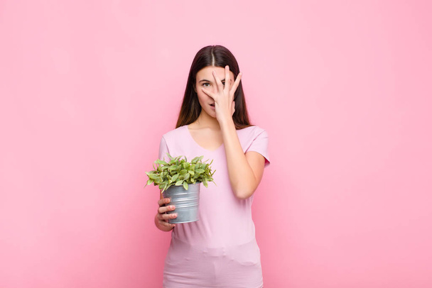 young pretty woman looking shocked, scared or terrified, covering face with hand and peeking between fingers with a plant - Фото, зображення