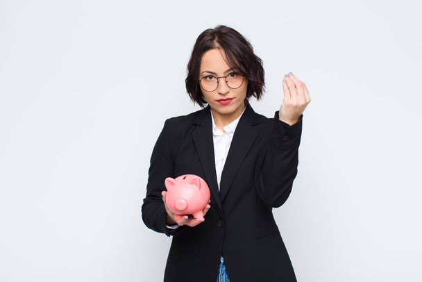 young pretty woman making capice or money gesture, telling you to pay your debts! with a piggy bank - Foto, afbeelding