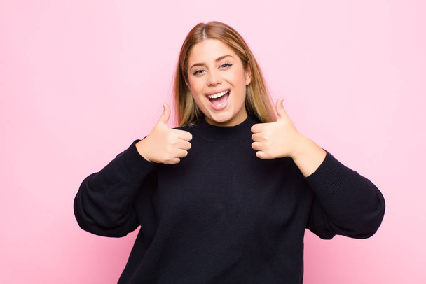 young blonde woman smiling broadly looking happy, positive, confident and successful, with both thumbs up against flat wall - 写真・画像