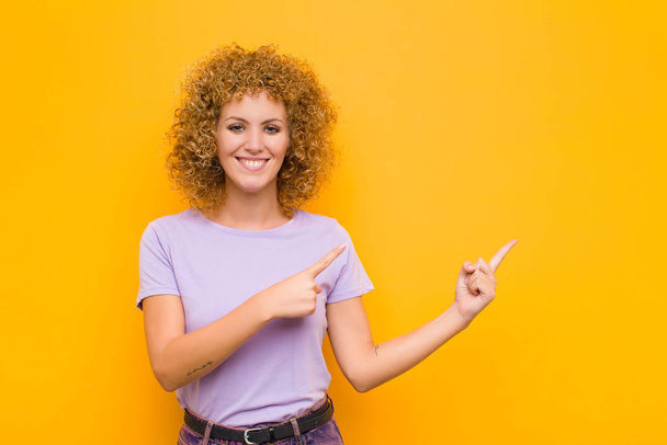 young afro woman smiling happily and pointing to side and upwards with both hands showing object in copy space against orange wall - Photo, Image