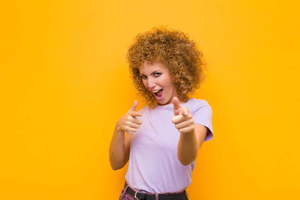 young afro woman feeling happy, cool, satisfied, relaxed and successful, pointing at camera, choosing you against orange wall - Foto, afbeelding