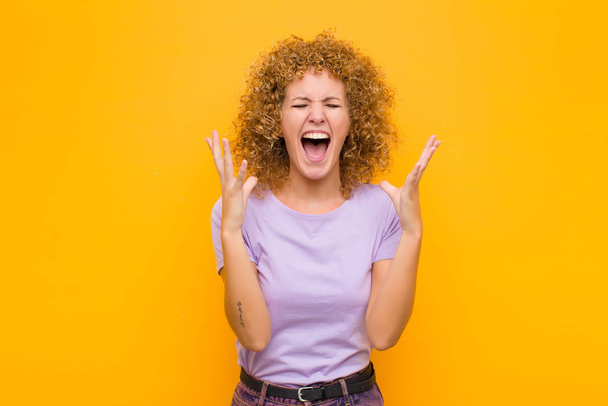 young afro woman furiously screaming, feeling stressed and annoyed with hands up in the air saying why me against orange wall - Fotoğraf, Görsel
