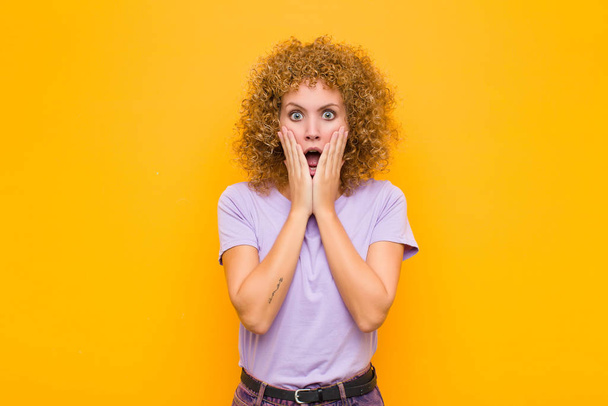 young afro woman feeling shocked and scared, looking terrified with open mouth and hands on cheeks against orange wall - 写真・画像