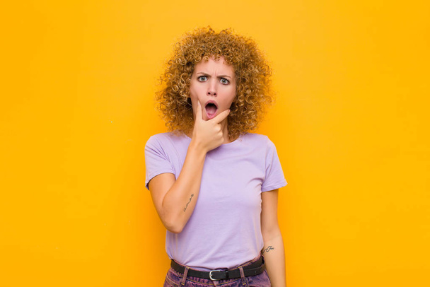 young afro woman with mouth and eyes wide open and hand on chin, feeling unpleasantly shocked, saying what or wow against orange wall - Foto, immagini