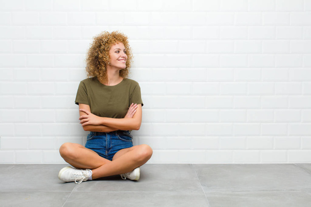 young afro woman smiling gleefully, feeling happy, satisfied and relaxed, with crossed arms and looking to the side sitting on a floor - Фото, изображение