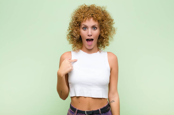 young afro woman looking shocked and surprised with mouth wide open, pointing to self against green wall - Photo, image