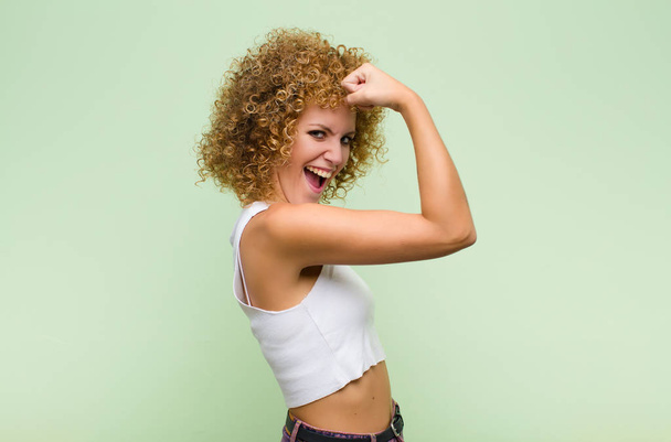 young afro woman feeling happy, satisfied and powerful, flexing fit and muscular biceps, looking strong after the gym against green wall - Photo, Image