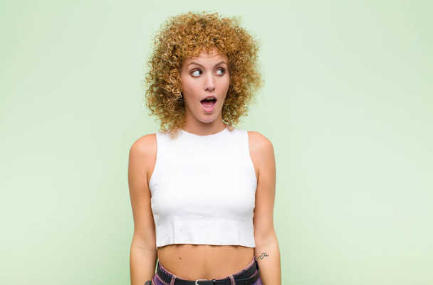 young afro woman feeling shocked, happy, amazed and surprised, looking to the side with open mouth against green wall - Φωτογραφία, εικόνα