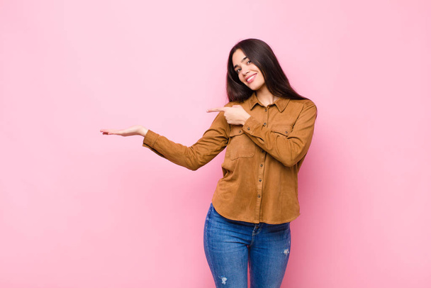 young pretty woman smiling, feeling happy, carefree and satisfied, pointing to concept or idea on copy space on the side against pink wall - 写真・画像
