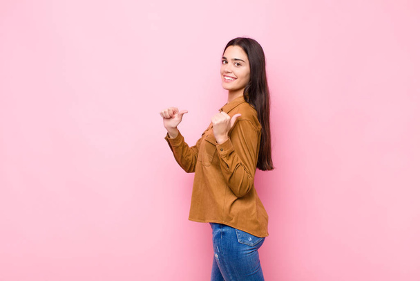 young pretty woman smiling cheerfully and casually pointing to copy space on the side, feeling happy and satisfied against pink wall - 写真・画像