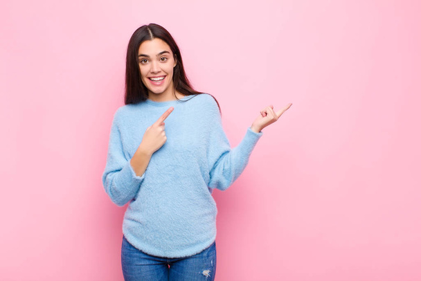 young pretty woman smiling happily and pointing to side and upwards with both hands showing object in copy space against pink wall - Zdjęcie, obraz