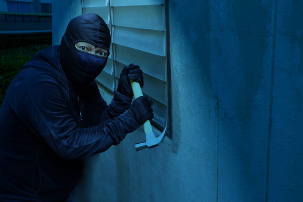A maszkos tolvaj kalapáccsal töri be az ablakot, hogy éjjel ellopja a házat, és érzékeli az otthoni biztonsági rendszerek kameráinak felvételeit.. - Fotó, kép