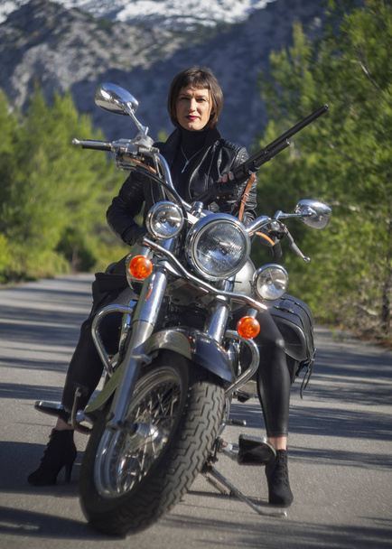 Una mujer en una chaqueta de cuero negro con un rifle de carabina en una motocicleta helicóptero en Grecia en una carretera en el bosque en las montañas
 - Foto, imagen