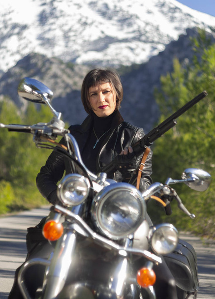 Siyah deri motorcu ceketli bir kadın. Karabina tüfeğiyle Yunanistan 'da dağlardaki ormanda bir yolda motosiklet kullanıyor. - Fotoğraf, Görsel