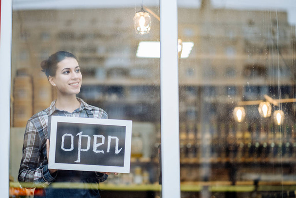 woman stands and opens a wide sign through the shop window and smile. - Photo, Image