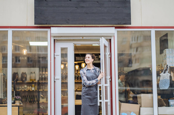 Портрет девушки-официантки в фартуке, стоящей в дверях кофейни и смотрящей на камеру и улыбающейся.
 - Фото, изображение