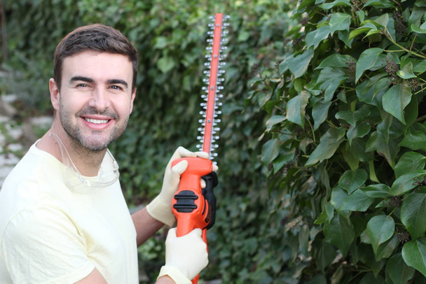 close-up portrait of handsome young man with electric saw cutting bushes in garden - Photo, Image