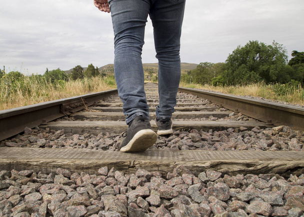 Pie joven irreconocible caminando sobre las vías del tren en una escena rural en un día nublado
 - Foto, Imagen