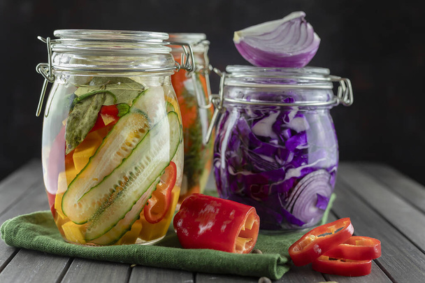 three glass jars of fermented red cabbage, cucumbers, chili pepper, vegetables on a dark background. using textile green. fermentation is a source of probiotic. popular vegetarian food. copy space - Photo, Image
