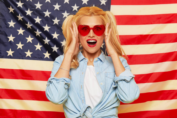 American girl. Happy young woman in heart shape sunglasses on USA flag  background. Funny and surprised human face. Patriotism concept - Фото, изображение