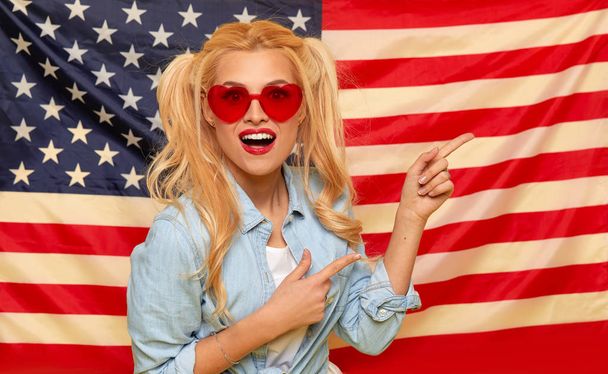 American girl. Happy young woman in heart shape sunglasses on USA flag  background. Funny and surprised human face. Patriotism concept - Φωτογραφία, εικόνα