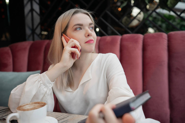 Νεαρή γυναίκα πίνει ζεστό καφέ και μιλάει στο τηλέφωνο στο καφέ κρατώντας σημειωματάριο - Φωτογραφία, εικόνα