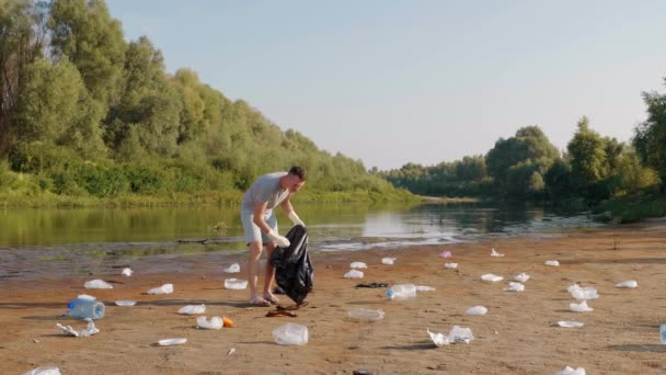 A man is collects plastic trash on the banks of a dry and polluted river or lake - Imágenes, Vídeo