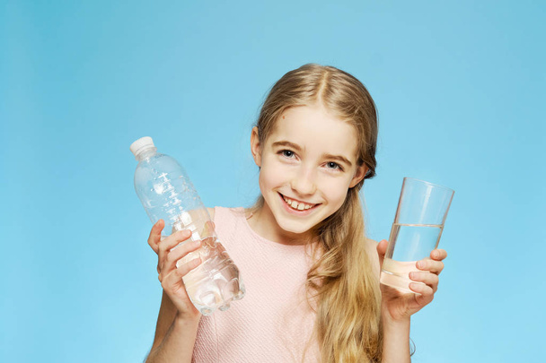 A little Caucasian girl holds a glass and a bottle of water in her hands. Drinking mode. - Foto, imagen