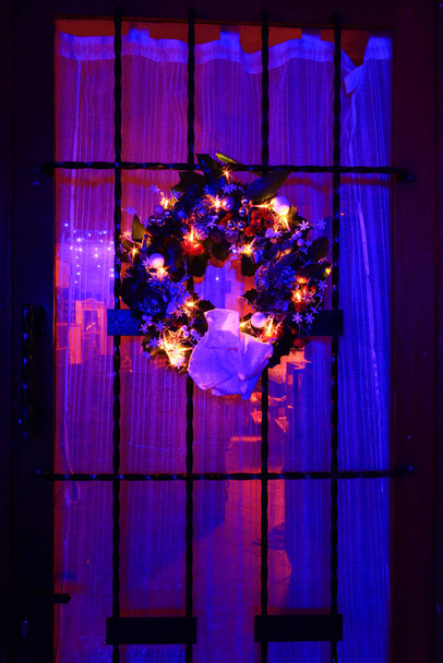 Corona de Adviento colgando de la puerta de una casa. Decoración navideña con una guirnalda ligera, con estrellas, muérdago, bolas y muñeco de nieve. Fondo bokeh colorido rojo y azul
. - Foto, imagen