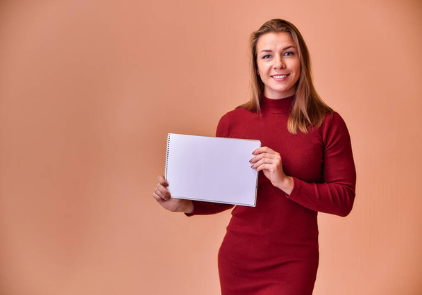 Portrait of a pretty blonde girl with a beautiful smile and excellent teeth in a burgundy dress with a folder in her hands on a pink background. Cute looks at the camera, standing straight. - Photo, Image