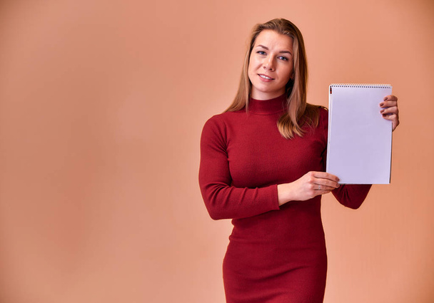 Portrait of a pretty blonde girl with a beautiful smile and excellent teeth in a burgundy dress with a folder in her hands on a pink background. Cute looks at the camera, standing straight. - Foto, Imagen