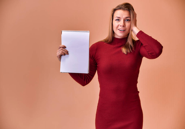 Portrait of a pretty blonde girl with a beautiful smile and excellent teeth in a burgundy dress with a folder in her hands on a pink background. Cute looks at the camera, standing straight. - Foto, Imagem