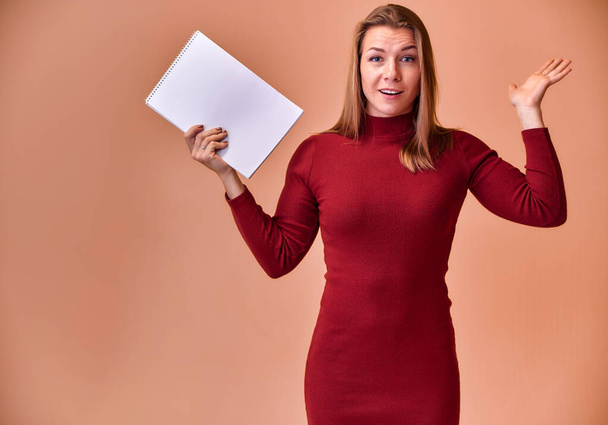 Portrait of a pretty blonde girl with a beautiful smile and excellent teeth in a burgundy dress with a folder in her hands on a pink background. Cute looks at the camera, standing straight. - Foto, immagini