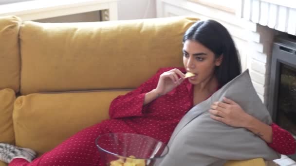 Persian woman at home watching TV eating chips potatoes - Filmati, video