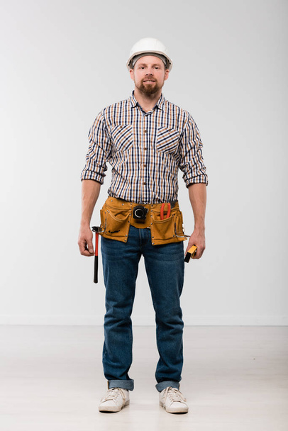 Young technician with toolbelt on his waist standing in front of camera and looking at you in isolation - Фото, зображення