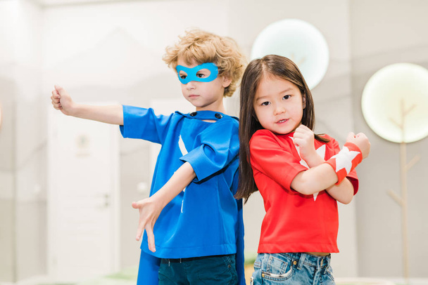Duas crianças interculturais em trajes de super-heróis de pé perto um do outro na frente da câmera durante o jogo
 - Foto, Imagem