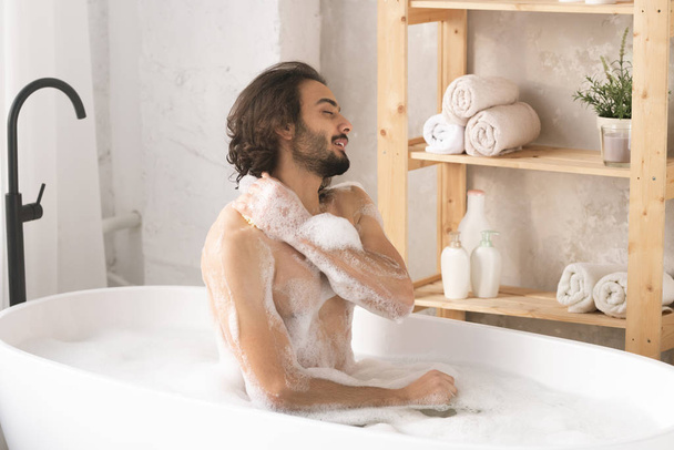 Молодий красивий голий чоловік сидить у ванні з гарячою водою і піною, миє тіло і насолоджується процедурою
 - Фото, зображення