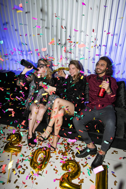 Młodzi radośni przyjaciele z piwem siedzący na miękkiej kanapie podczas zabawy podczas noworocznej imprezy w klubie nocnym - Zdjęcie, obraz