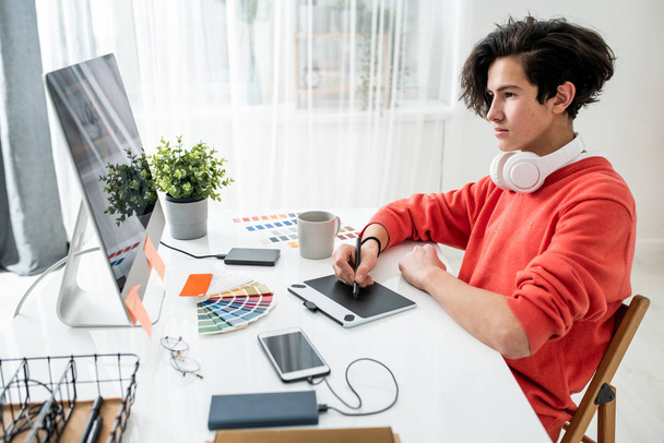 junger Mann mit Kopfhörern schaut auf Computerbildschirm, während er Grafik-Tablet und Stift benutzt, um Fotos zu retuschieren - Foto, Bild