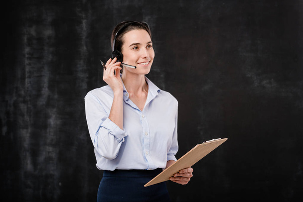 Cheerful young secretary or operator with clipboard talking to clients in microphone over black background - Fotoğraf, Görsel